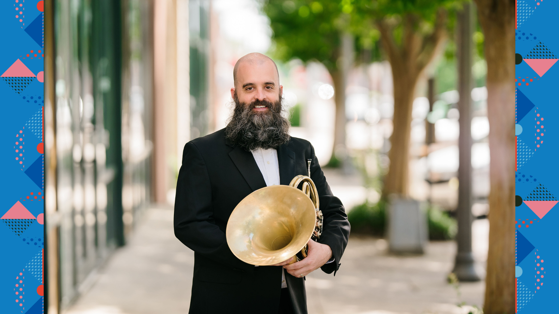 Rob Detjen in a suit holding a french horn outside on the sidewalk with trees and a building in the background.